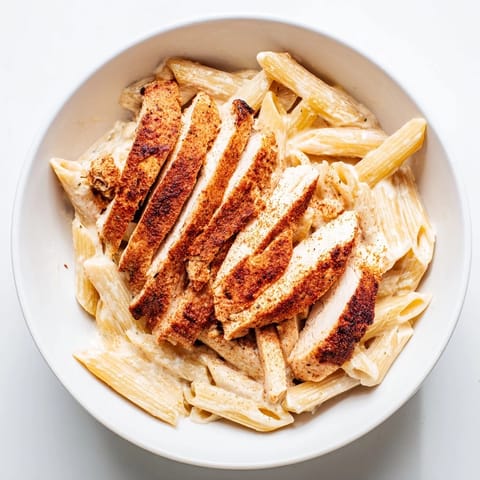 A vibrant bowl of Spicy Chicken Caesar Pasta features golden penne, parmesan shavings, and crunchy croutons on a white marble counter.  